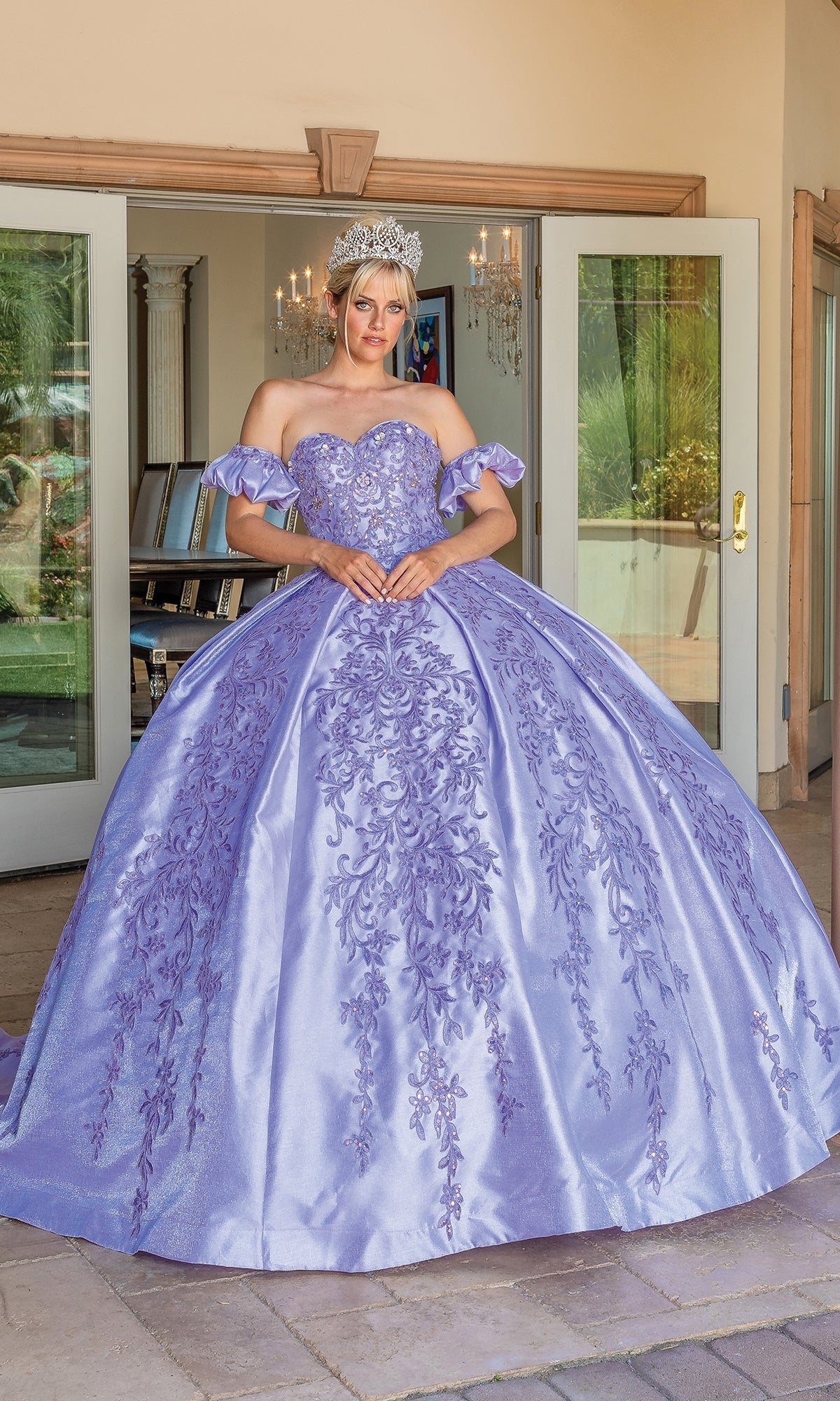 A woman wearing a silver crown and the Quinceanera Dress 1644 By Dancing Queen, a lavender strapless ball gown with floral embroidery, stands in front of open glass doors.
